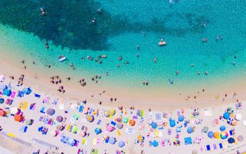 People bathing in the sun at the beach, aerial view with clear blue water and sandy beach. Corfu island Kerkyra, Greece (photo via CalinStan/iStock/Getty Images Plus)