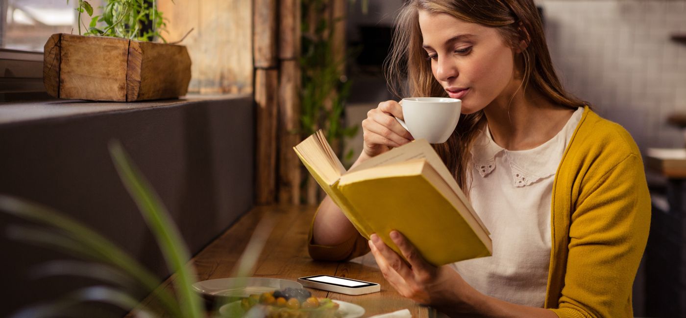 Image: A woman sits at a cafe and reads with a warm drink. (Photo Credit: WavebreakMediaMicro / AdobeStock)