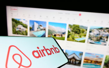 Airbnb logo displayed on a smartphone in front of a listings webpage.